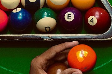 A hand holds an orange billiard ball with a yellow TYPO3 logo, over a pool table. In the background, striped and solid balls in a rack spell out IDEA with each ball featuring a letter.